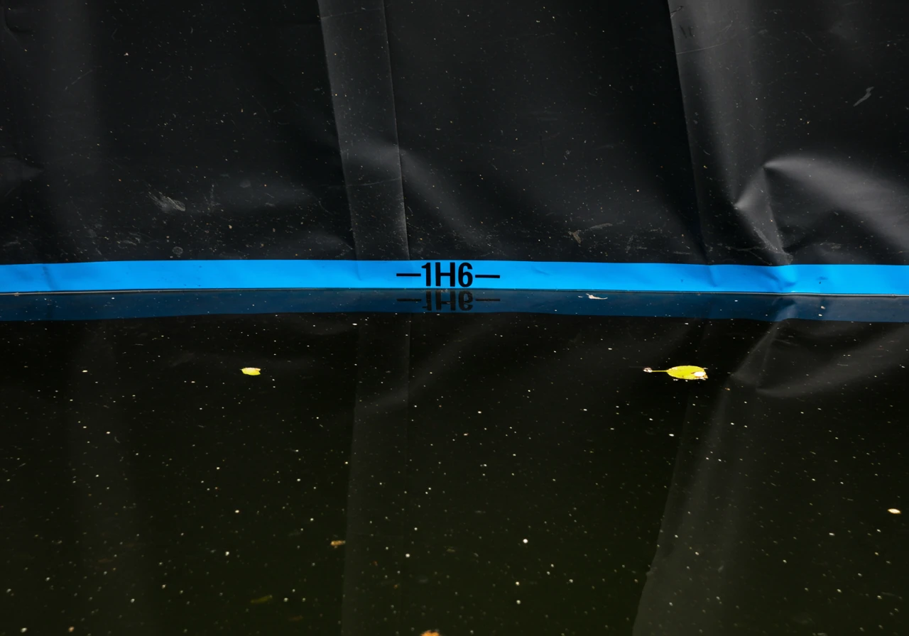 An eye-level shot from inside a calm pond, looking towards the liner edge. A distinct mark, like a piece of bright blue waterproof tape or a clear grease pencil line, is visible on the dark liner wall. The water level is clearly shown about one inch below this mark, visually demonstrating the water drop during a leak test. The water surface is still with minimal reflection.